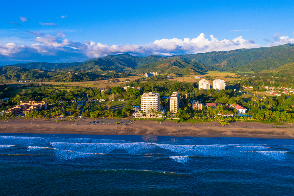 Aerial view of Jaco Beach, Costa Rica, with vibrant coastline, modern amenities, and lush mountains, highlighting the lifestyle, schools, healthcare, and entertainment options promoted by Jaco Coldwell Banker.