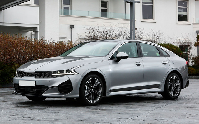 KIA K5 – передне- или полноприводный седан бизнес-класса, позиционирующийся в качестве так называемого «четырехдверного купе», который может похвастать смелым дизайном с ярким деталями, современным и вместительным салоном, в меру производительными моторами и широким перечнем прогрессивных опций, да еще и за «относительно небольшие деньги»… Этот «кореец» адресован, в первую очередь, молодой целевой аудитории, для которой важно, как автомобиль выглядит и едет, однако только лишь этими рамками его «потенциальные покупатели» не ограничиваются… Официальная презентация KIA K5 пятого воплощения (предыдущее поколение этого седана в России известно как Optima) состоялась 21 ноября 2019 года на специальном мероприятии в Сеуле, а уже 12 декабря начались продажи трехобъемника на рынке Южной Кореи. После очередной «реинкарнации» автомобиль пережил смену дизайнерской концепции, превратившись в «седан в стиле фастбэка», укрупнился в размерах, превзойдя одного из лидеров класса в лице Toyota Camry, «переехал» на новую платформу, впервые в своей истории получив полноприводную трансмиссию, «вооружился» новыми двигателями и обзавелся большим количеством прогрессивных «фишек».Внешне «fifth K5» может похвастать смелым, элегантным, по-спортивному подтянутым и динамичным обликом, в котором хватает ярких дизайнерских решений. Анфас четырехдверка выставляет напоказ нахмуренные фары сложносочиненной формы с зигзагами ходовых огней, как бы интегрированные в «нос тигра» радиаторной решетки с крупноячеистой структурой, и агрессивный бампер, а его выразительную корму украшают стильные фонари, объединенные в сплошную линию, и мощный бампер с двумя трапециевидными патрубками выхлопной системы.Но наиболее выигрышно автомобиль выглядит в профиль, поскольку «щеголяет» стремительным и приземистым силуэтом а-ля «четырехдверное купе» с длинным капотом, покатой линией крыши, сильно заваленным задним стеклом и коротеньким «хвостиком» багажника, основательности которому добавляют колоритные боковины и внушительные росчерки колесных арок. Размеры и вес По своим габаритам KIA K5 2020 модельного года относится к E-сегменту по европейским меркам: в длину седан достигает 4905 мм, из которых на 2850 мм простирается межосевое расстояние, а в ширину и в высоту насчитывает 1860 мм и 1445 мм соответственно. В снаряженном состоянии масса четырехдверки варьируется от 1410 до 1515 кг, в зависимости от модификации. Интерьер Внутри «Оптима» пятой генерации отличается элегантным, современным и весьма солидным интерьером, отвечающим всем тенденциям цифровой эры.На рабочем месте водителя располагаются трехспицевый мульти-руль с рельефным ободом, слегка усеченным в нижней части, и полностью виртуальные приборы, «нарисованные» на дисплее диагональю 12.3 дюйма (правда, у начальных вариантов – аналоговые шкалы да скромный 4.2-дюймовый экран). Изысканную центральную консоль возглавляет 10.25-дюймовый тачскрин, под которым базируются асимметричные дефлекторы вентиляции и лаконичный «пульт» климатической установки с сенсорным управлением. Убранство автомобиля отделано преимущественно добротными материалами отделки, причем «топовые» исполнения способны похвастать поверхностями с эффектом натурального дерева или металла.Салон «пятого» KIA K5 обладает пятиместной компоновкой, причем на достаточный запас свободного пространства могут рассчитывать обитатели обоих рядов сидений. На передних местах находятся эргономично спрофилированные кресла с отчетливым боковым профилем и широкими диапазонами регулировок, которые в «топовых» версиях дополняются электроприводом, подогревом и вентиляцией. На втором ряду – комфортабельный диван с откидным центральным подлокотником и подогревом, а также собственные дефлекторы вентиляции.На втором ряду – комфортабельный диван с откидным центральным подлокотником и подогревом, а также собственные дефлекторы вентиляции.Второй ряд сидений складывается несколькими частями, но ровного пола в таком случае не получается. В нише под фальшполом – полноценная «запаска» и набор инструментов, аккуратно уложенный в органайзер. Технические характеристики Для Kia K5 fifth-генерации на российском рынке предлагается два четырехцилиндровых бензиновых мотора: Штатно автомобиль оснащается 2.0-литровым «атмосферником» MPI с распределенным впрыском, 16-клапанным ГРМ типа DOHC и регулируемыми фазами газораспределения, генерирующим 150 лошадиных сил при 6200 об/минуту и 192 Нм крутящего момента при 4000 об/минуту. Более производительным версиям полагается атмосферный двигатель GDI рабочим объемом 2.5 литра с системой непосредственного впрыска, 16-клапанным ГРМ типа DOHC и фазовращателями на впуске и выпуске, который выдает 194 л.с. при 6100 об/минуту и 246 Нм пиковой тяги при 4000 об/минуту. Оба мотора сочетаются исключительно с ведущими колесами передней оси и гидромеханическими автоматическими коробками передач, но первый вариант – с 6-диапазонной, а второй – с 8-ступенчатой.
С места до 100 км/ч четырехдверка ускоряется через 8.6-10.6 секунды, а ее «максималка» укладывается в 200-210 км/ч. Расход топлива у седана варьируется от 7.1 до 7.2 литров на каждую «сотню» пробега в комбинированном цикле в зависимости от модификации. Конструктивные особенности Пятый «релиз» KIA K5 базируется на «переднеприводной» архитектуре с поперечно расположенным силовым агрегатом и цельнометаллическим несущим кузовом, на широкую долю состоящим из стали высокопрочных сортов. Седан комплектуется независимыми подвесками со стальными пружинами, пассивными амортизаторами и поперечными стабилизаторами «по кругу»: на передней оси – архитектурой типа МакФерсон, на задней – многорычажной системой. Стандартно автомобиль обладает рулевым механизмом типа «шестерня-рейка» с интегрированным электрическим усилителем управления. И спереди, и сзади четырехдверка оснащается дисковыми тормозными устройствами (но в первом случае – вентилируемыми), дополненными ABS, EBD и BAS.