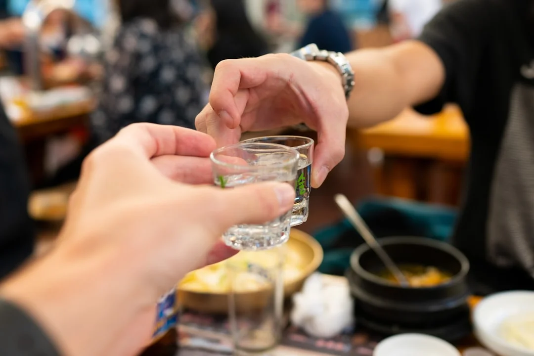 Two people sharing a soju shot glass at a Korean restaurant, with traditional Korean side dishes visible in the background