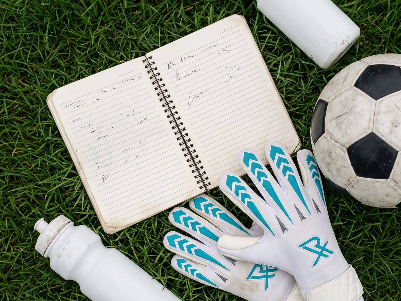 A notebook open on the grass beside a football, a pair of goalkeeper gloves and a water bottle