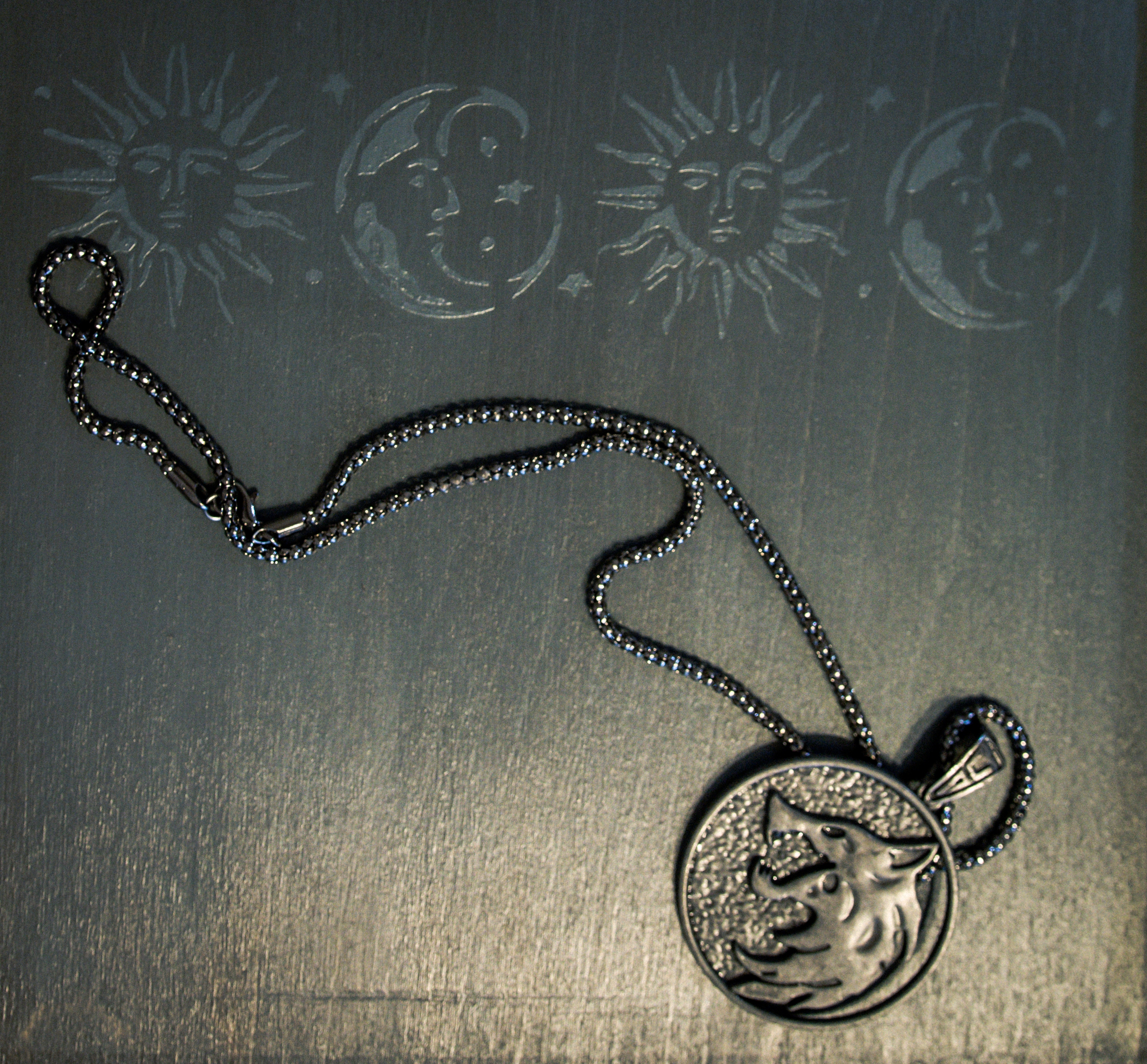 A metal medallion with a wolf's head, like Geralt's in the Netflix series, rests on a black wooden surface. It gleams in the light. Above it, painted on the wood, are images of alternating suns and moons.