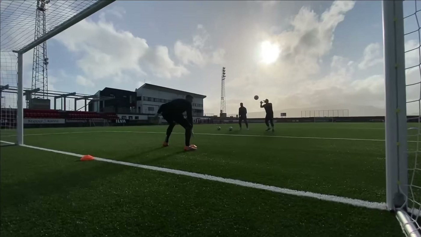 Goalkeeper training alone under floodlights at night with a few cones set out on the pitch