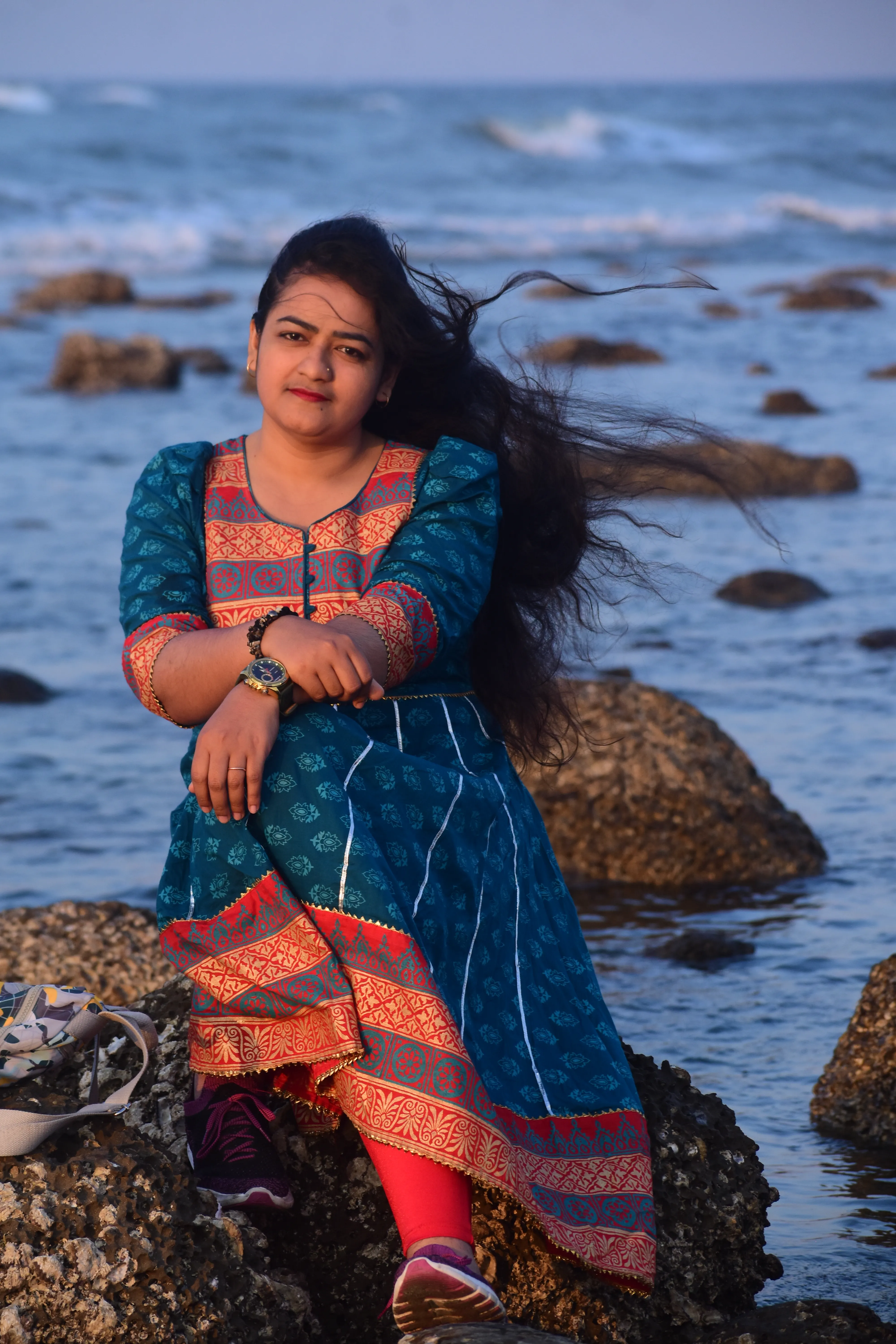 Elizabeth A young woman in a teal and red traditional outfit sitting on rocky ocean shores with long hair blowing in the wind.