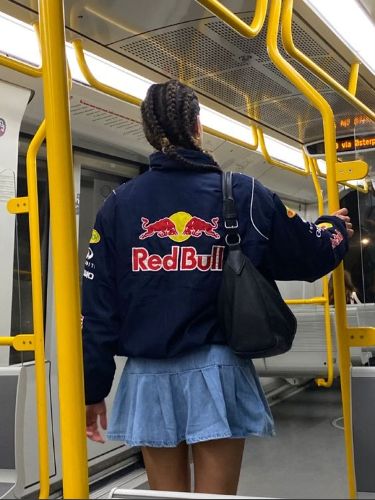 A photo of a woman in a metro wearing a Red Bull Racing jacket and a mini denim skirt. The photo is taken from the back.