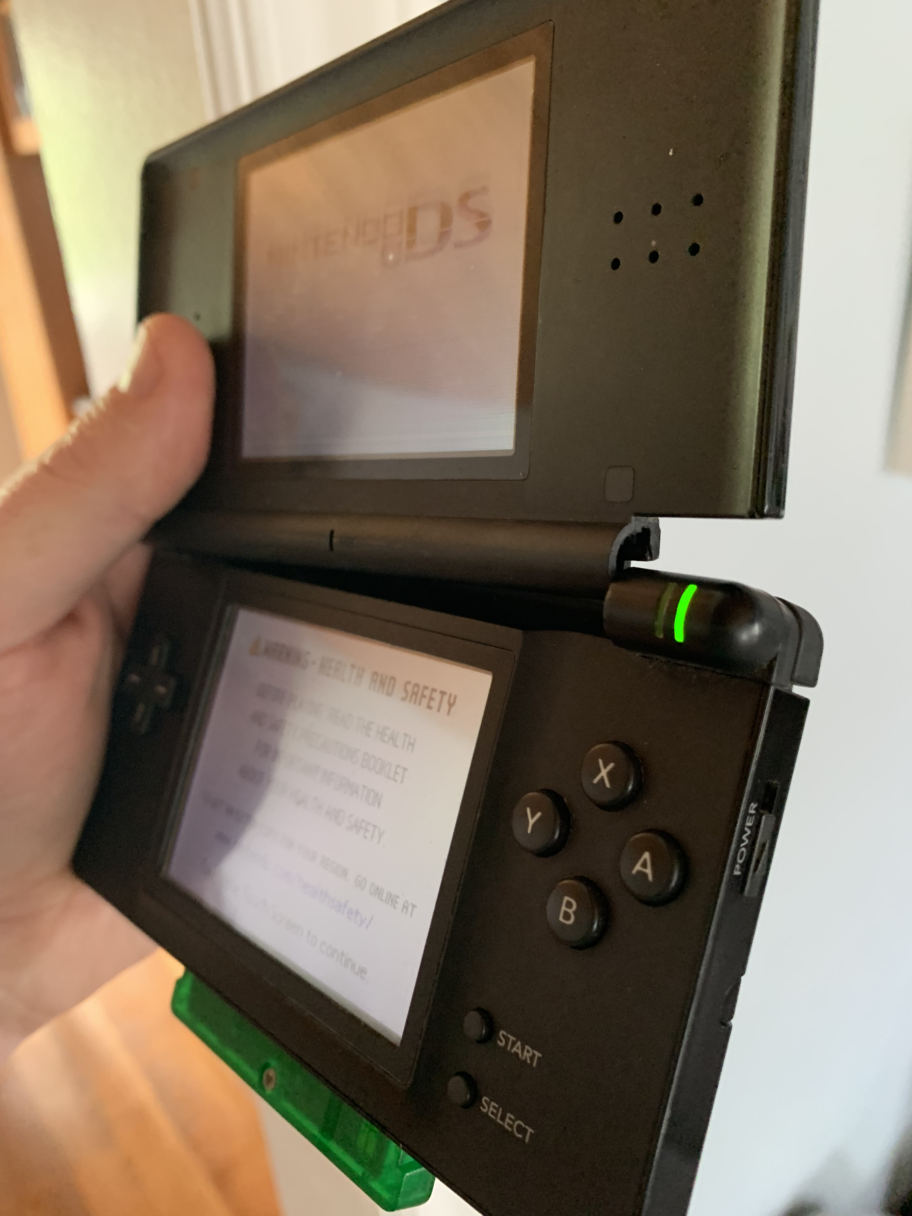 A black DS Lite with a green GBA cartridge sticking out, and the screen is broken off of the right side. The screen is displaying the DS startup screen.