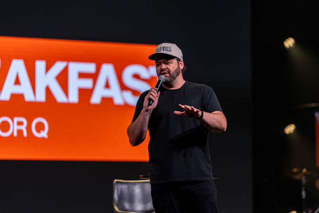 Will Jackson speaking on stage, holding a microphone and gesturing toward the audience