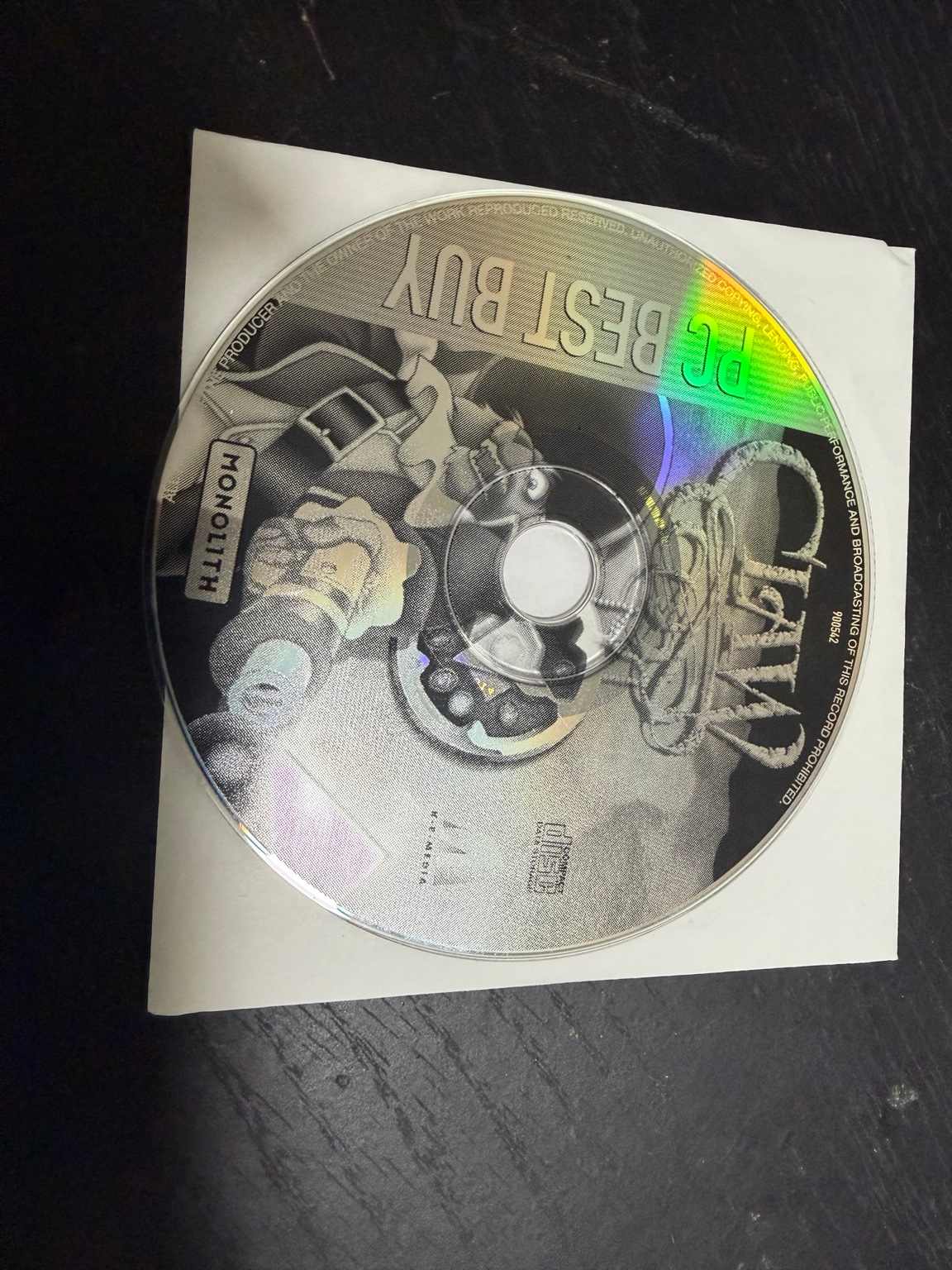 A loose PC Best Buy Claw CD with Monolith branding resting on a paper sleeve.