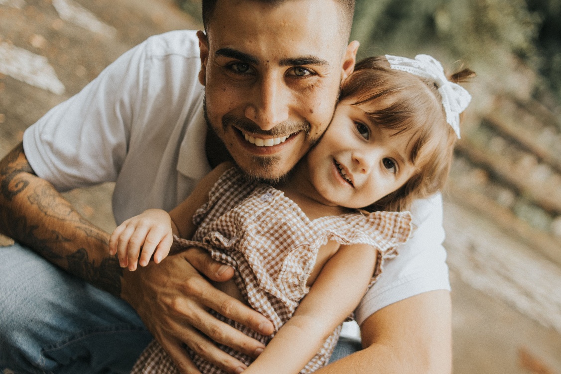 Smiling man hugging a child outdoors, both leaning close together.