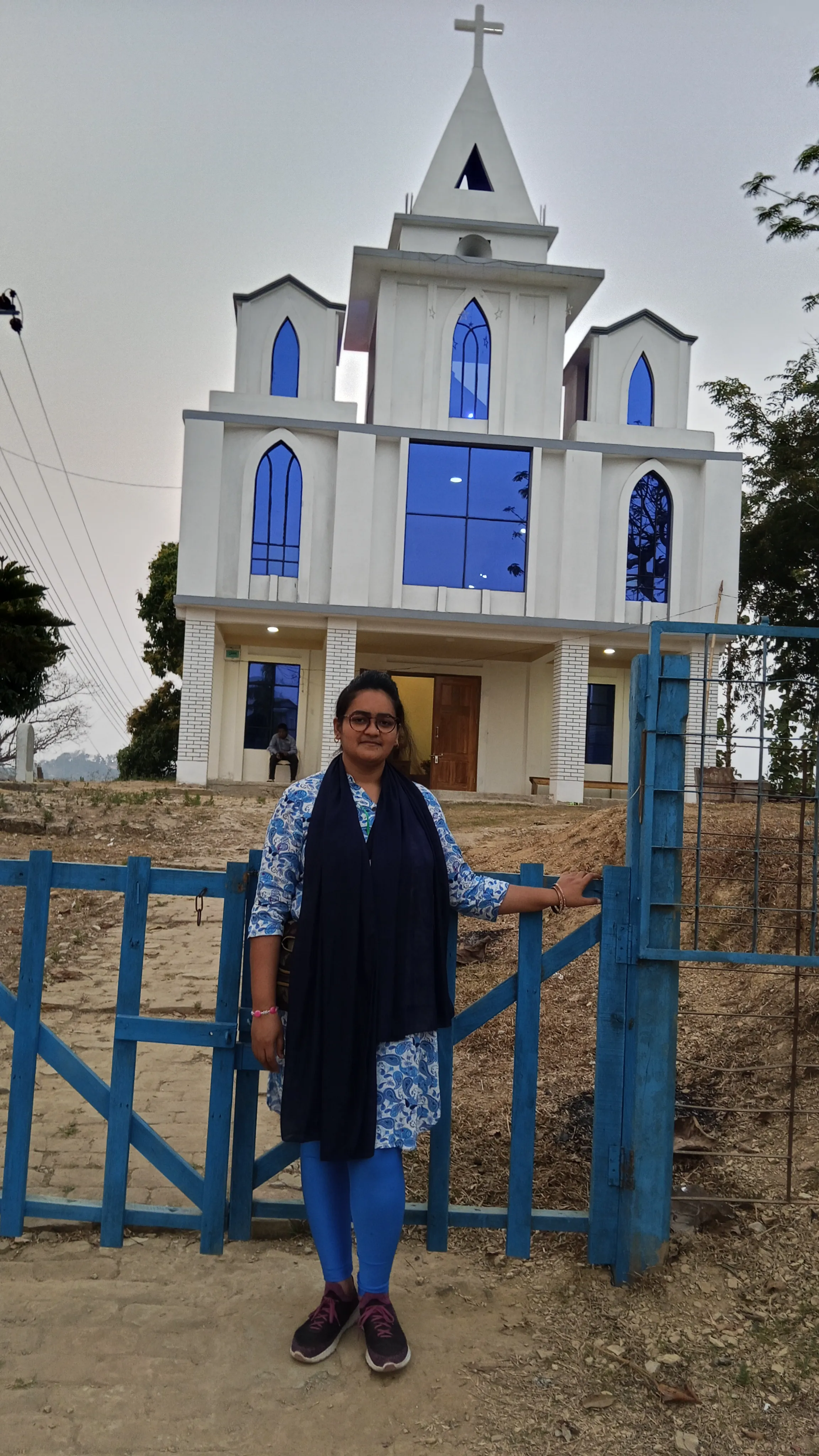 Elizabeth A woman wearing glasses and a blue tunic standing outside a blue wooden fence with a tall white church in the background.