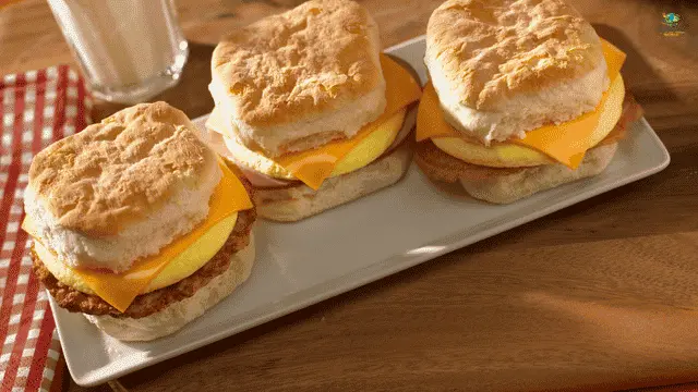 healthy freezer breakfast sandwiches A tray filled with halved ciabatta breakfast sandwiches stuffed with fresh arugula, egg, roasted red peppers, and leafy greens, with a hand reaching for one.
