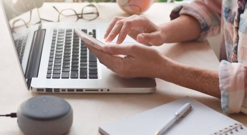 Mãos masculinas teclam um celular em frente a um notebook.