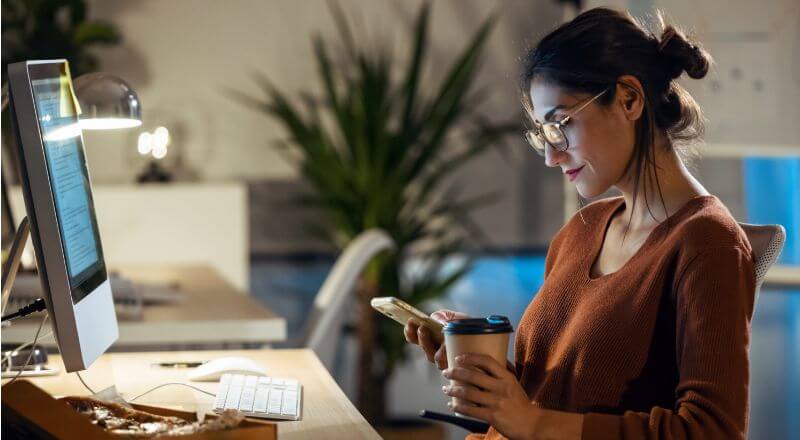 Mulher mexendo no celular e observando um notebook.