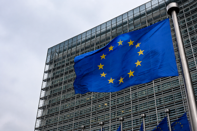 Photorealistic photojournalistic image of the European Union flag flying outside a modern glass government building in Brussels, overcast da