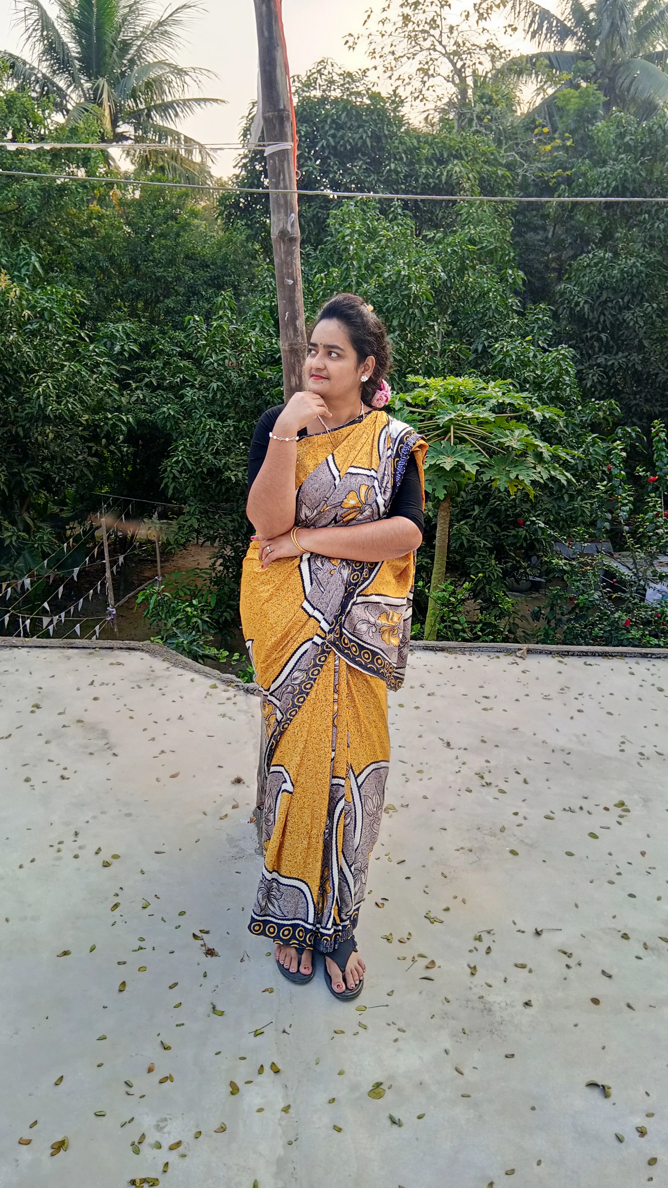 Elizabeth A woman wearing a yellow and patterned saree posing thoughtfully outdoors against a background of green foliage.