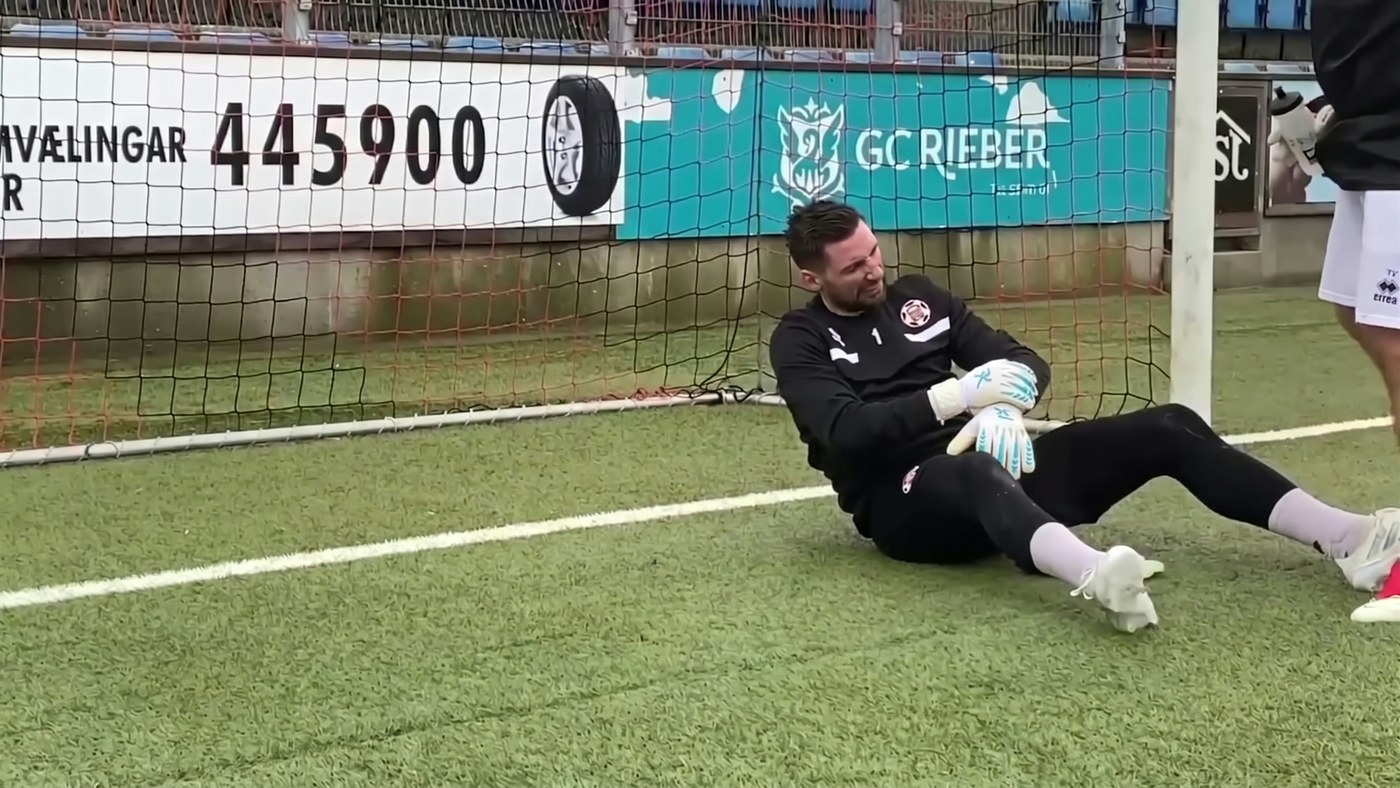Goalkeeper sat on the turf inside his goal after a save, checking his glove and wrist while play waits