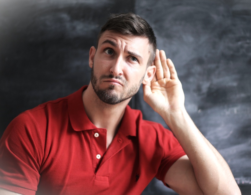 Man in a red polo shirt cupping his ear, listening with a puzzled expression against a chalkboard background