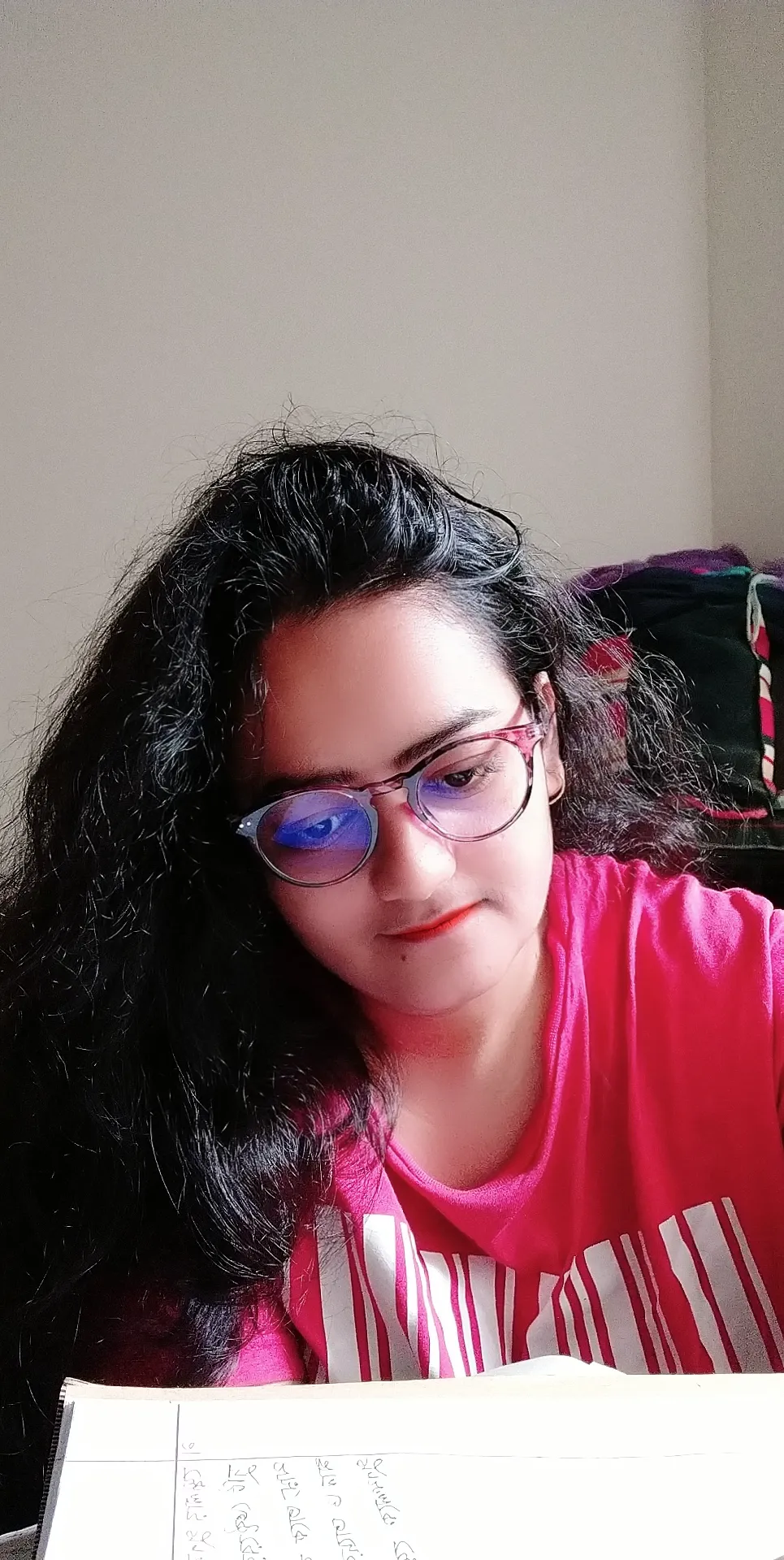 Elizabeth A young woman with long dark wavy hair wearing eyeglasses and a pink t-shirt looking down at a notebook while studying indoors.