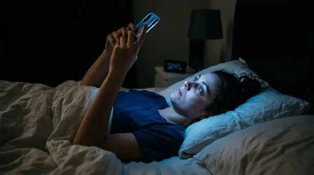 Person lying in dark bedroom with face illuminated by smartphone blue glow, sleepless and tired