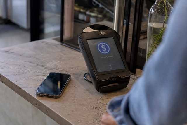 Photorealistic photojournalistic image of a modern retail counter with a smartphone and contactless payment terminal showing a glowing dolla