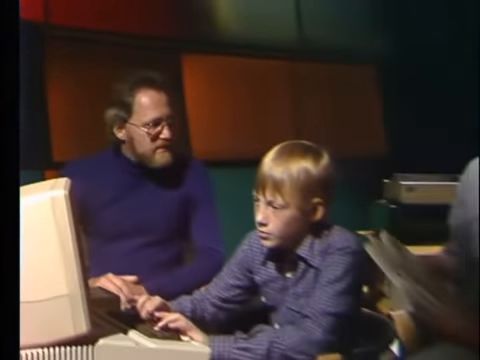 A child and two adults gathered around a computer during a television studio demonstration