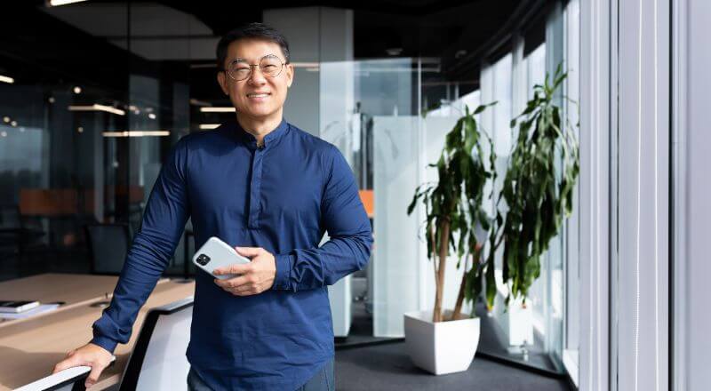 Homem asiático sorrindo e olhando para frente com um celular na mão.