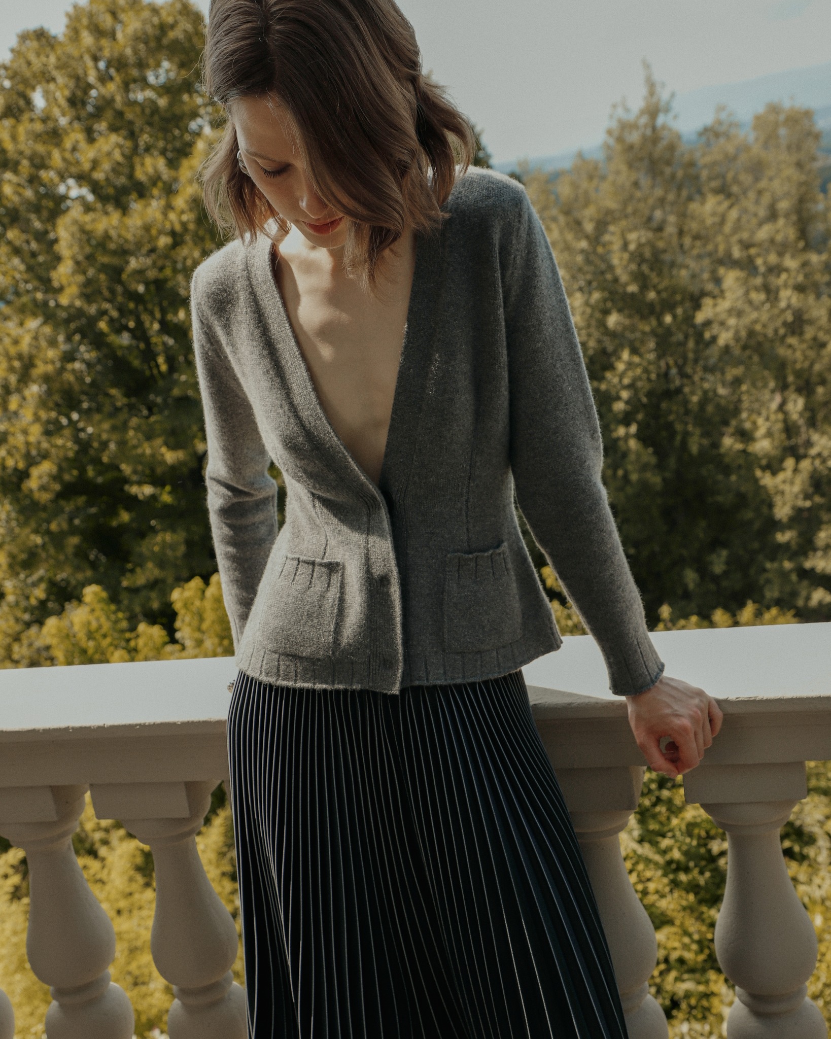 Malo Rituals: cardigan di cashmere e gonna plissettata Una donna per la campagna Malo Rituals Autunno Inverno 2025. Indossa un cardigan di cashmere grigio e una lunga gonna plissettata scura, appoggiata a un balcone. L'immagine evoca un'atmosfera di eleganza e serenità.