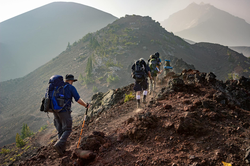 Group of hikers with full backpacks and trekking poles ascending a rocky volcanic mountain ridge under hazy mountain skies, illustrating essential pack gear for Hallasan hiking