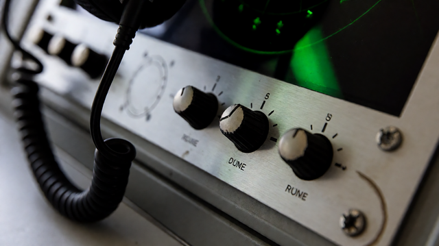 Close-up detail of an air traffic control console bezel with worn volume knobs, coiled black headset cable, green radar glow on brushed meta