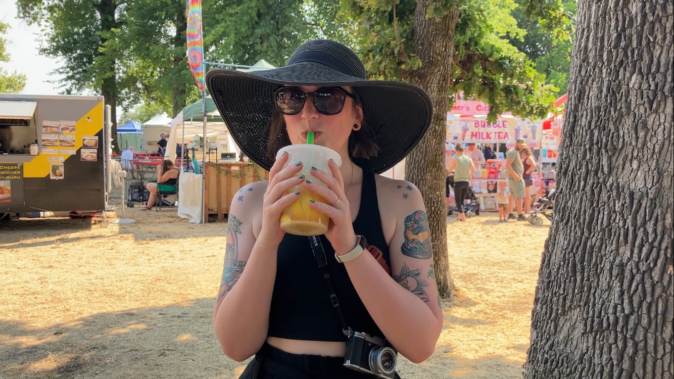 a white woman wearing a large black straw sunhat, drinking lemonade with boba from a to-go soup container the size of her head