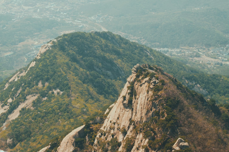 Dramatic rocky mountain ridgeline with volcanic granite spires rising above dense forest on a Korean mountain trail, with city skyline visible far below in the valley