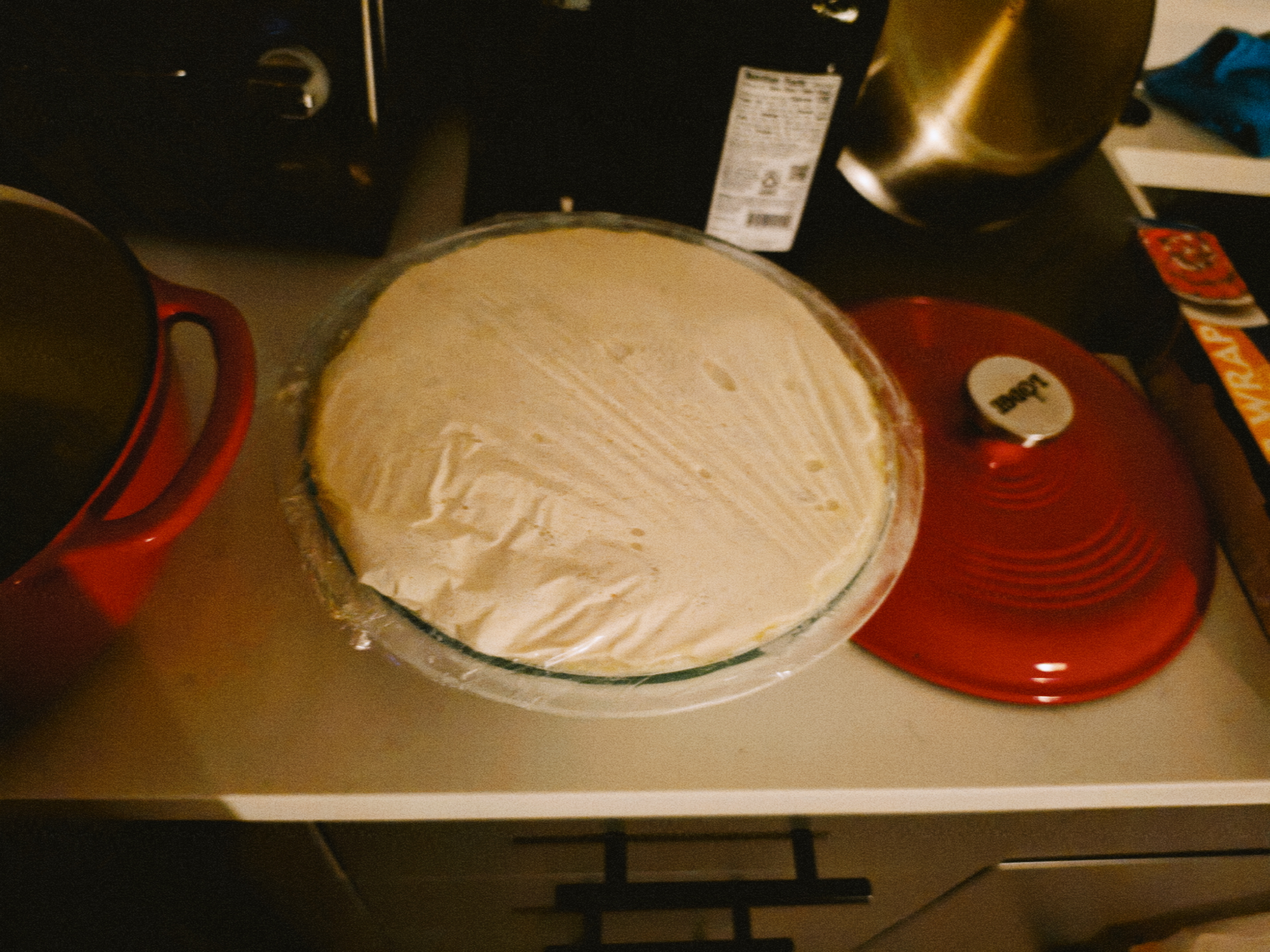 bigger and proofed dough in a glass bowl covered in plastic wrap. it looks like it is about to overflow.