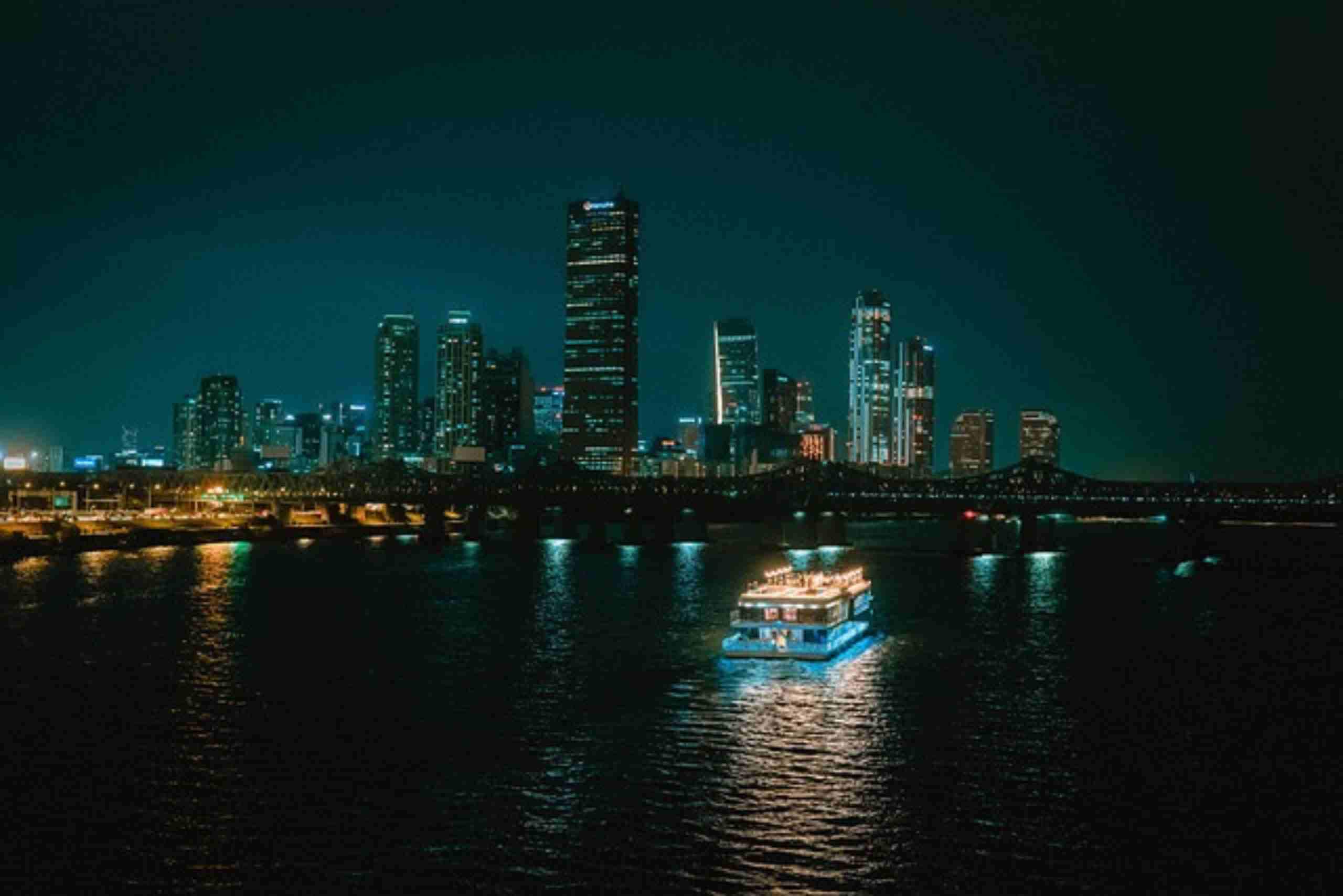  An image showing a night view of the Han River in Seoul, featuring a cruise boat and the city skyline.