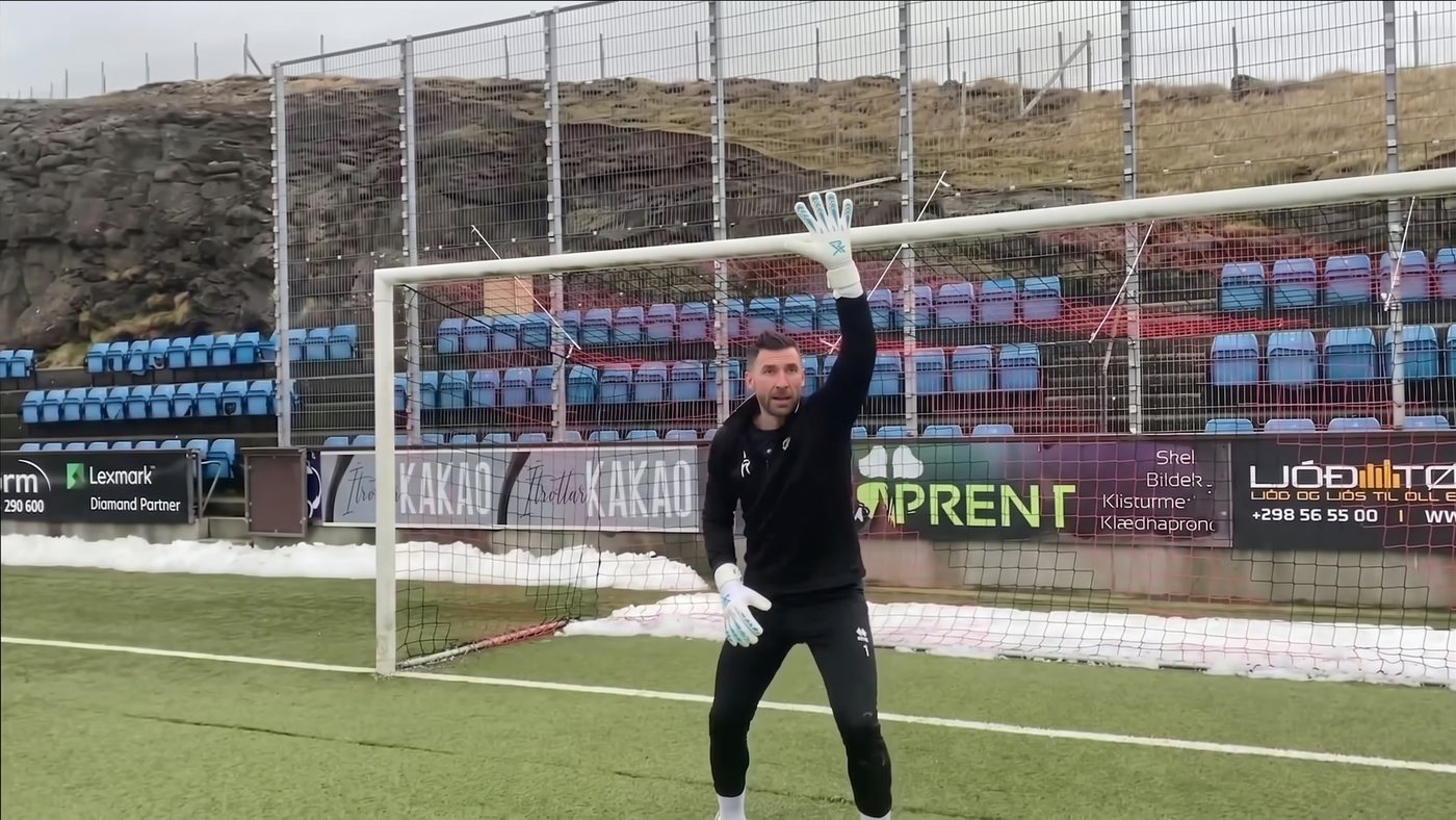 Goalkeeper in his goal with snow banked along the side of the pitch, one arm raised, shouting a single call to his defence