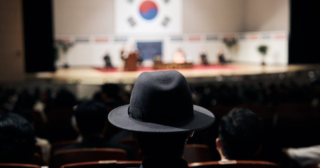 영화 암살자(들) 스틸컷, 태극기가 걸린 기념식장을 객석에서 바라보는 인물의 뒷모습