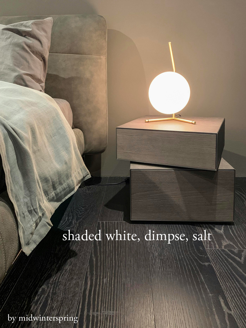 A photograph of a round bright light on an end table next to the corner of a bed. All of the colors in the room are shades of beige and the floor is grey-beige woodgrain