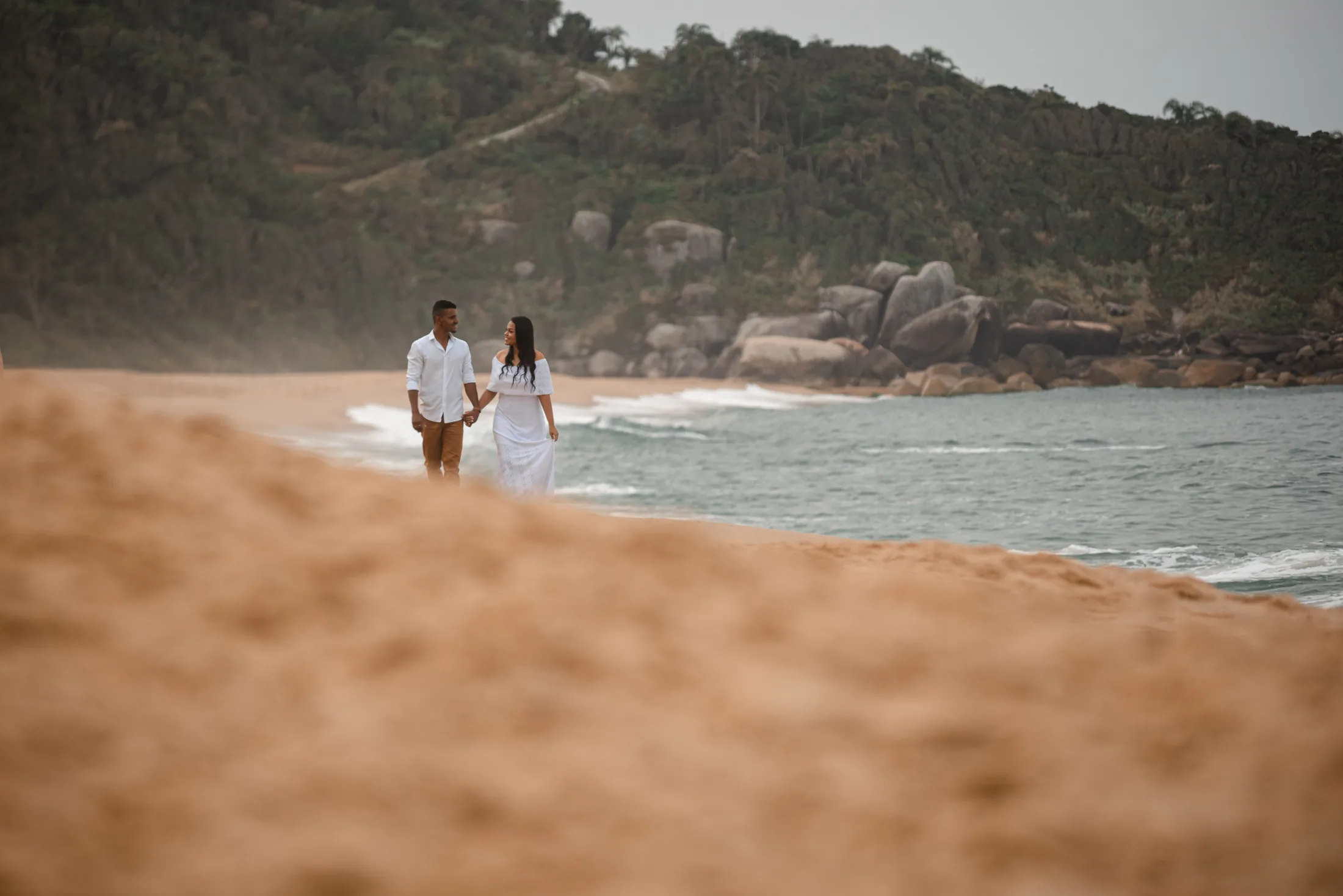 Casal caminhando na Praia de Taquaras com areia, mar e costão ao fundo em Balneário Camboriú