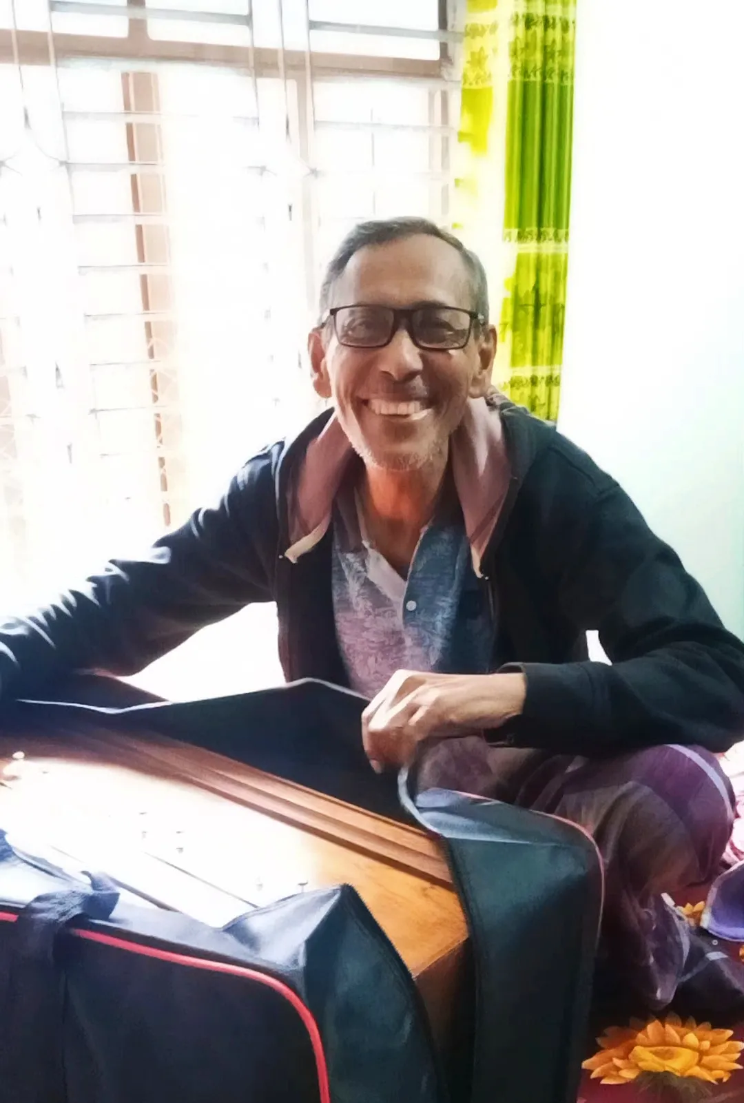 Elizabeth A smiling senior man wearing glasses unpacking a wooden harmonium from a black carrying case indoors.